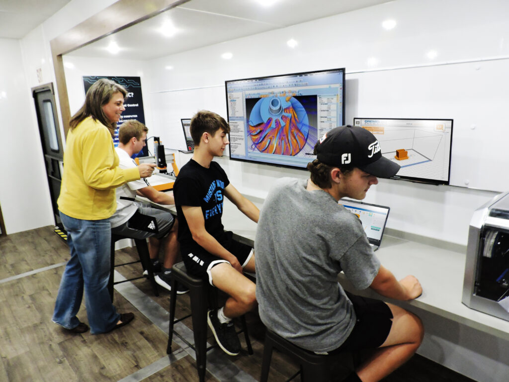Three individuals working on computers in a room with a large screen, part of the Tallapoosa CNC Students Mobile STEM Units initiative.